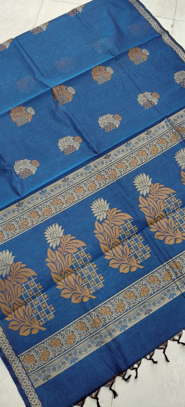 A blue negamam cotton saree with beige and gold block-printed floral motifs and a wide decorative border, laid flat on a light tiled floor.